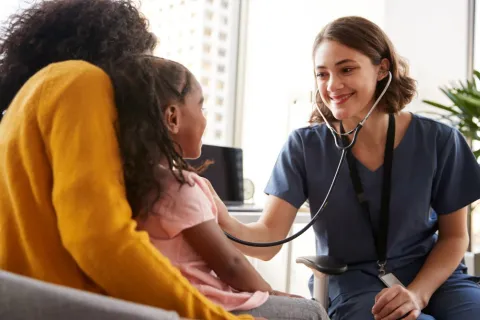 A smiling nurse practitioner listens to a young patient’s heart using a stethoscope.